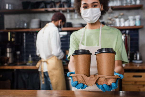Paper cups in hands of african american barista in medical mask on blurred background  - Photo, Image