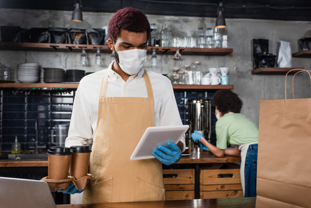African american barista in medical mask holding coffee to go and using digital tablet  - Photo, Image