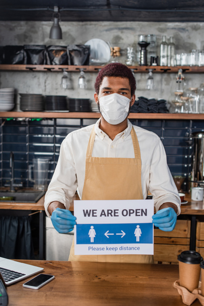 African american barista in medical mask and latex gloves holding signboard with we are open lettering near coffee in cafe  - Photo, Image