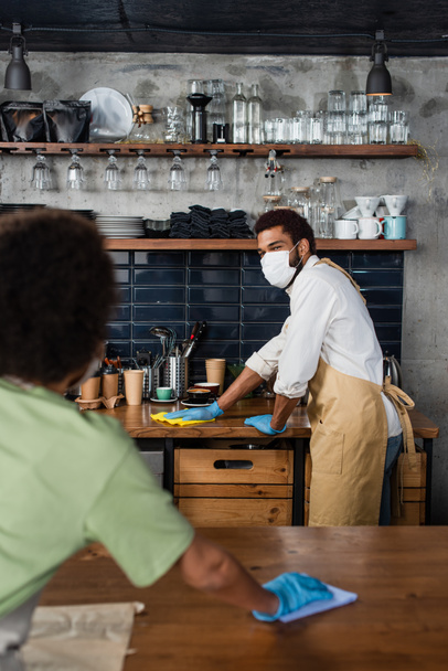 Barista afroamericano en máscara médica y guantes de látex limpiando encimera con trapo cerca de colega borroso  - Foto, Imagen