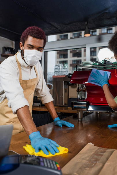 African american barista in medical mask cleaning bar near colleague in cafe  - Fotoğraf, Görsel