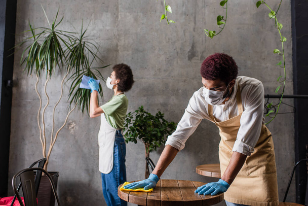 Afrikaanse amerikaanse barista in schort en medisch masker schoonmaken tafel in de buurt van collega op wazig achtergrond  - Foto, afbeelding