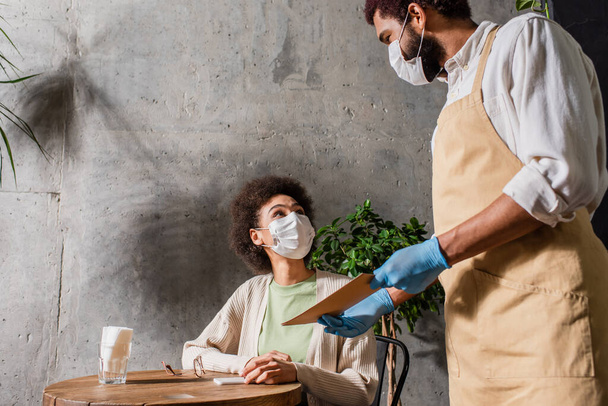 African american waiter in medical mask holding menu near client in cafe  - Photo, Image