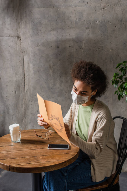 Cliente afroamericano en máscara médica mirando el menú cerca de anteojos y teléfonos inteligentes en la cafetería  - Foto, imagen