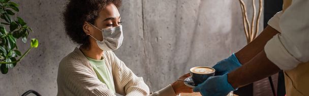医療マスクのアフリカ系アメリカ人女性は、ラテックス手袋、バナーでウェイターからコーヒーを取ります  - 写真・画像