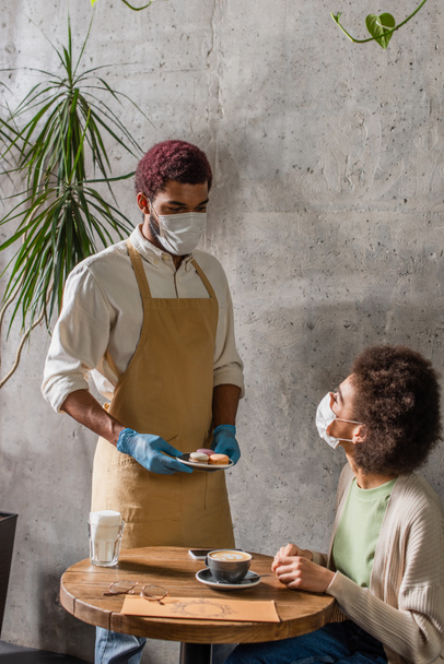 African american waiter in latex gloves holding macaroons near client in medical mask in cafe  - Photo, Image