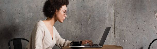 Vue latérale d'un pigiste afro-américain utilisant un ordinateur portable près d'une tasse dans un café, une bannière  - Photo, image