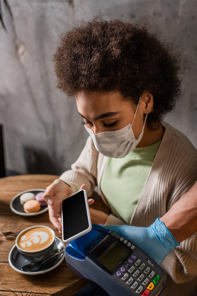 African american client in medical mask paying with smartphone near waiter with terminal in cafe  - Foto, Imagem