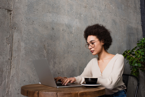 Giovane freelance afro-americano che utilizza il computer portatile vicino a una tazza di caffè nel caffè  - Foto, immagini