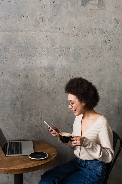 Vista lateral da mulher americana africana positiva com café usando smartphone perto do laptop na mesa no café  - Foto, Imagem