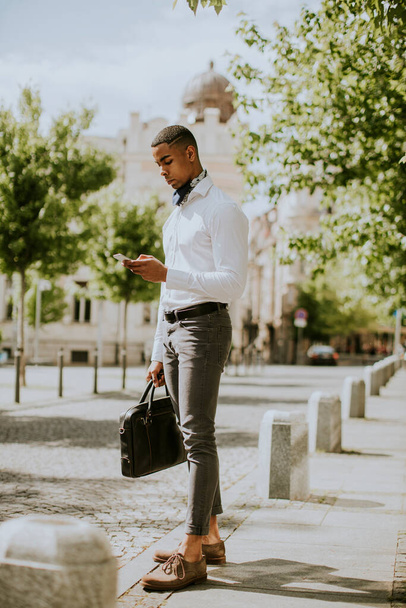 Bonito jovem empresário afro-americano usando um telefone celular enquanto espera por um táxi em uma rua - Foto, Imagem