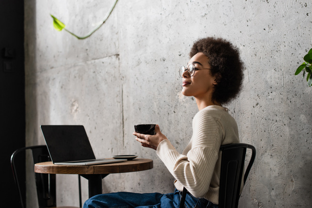 African american woman holding cup of coffee near laptop in cafe  - Photo, Image