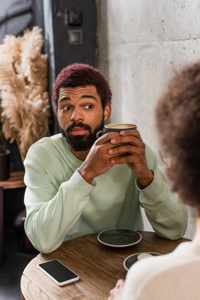 Amazed african american man with coffee looing at blurred girlfriend near smartphone in cafe  - Foto, imagen
