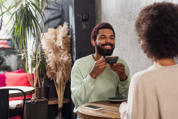 Positive african american man with coffee looking at girlfriend on blurred foreground in cafe  - Foto, imagen
