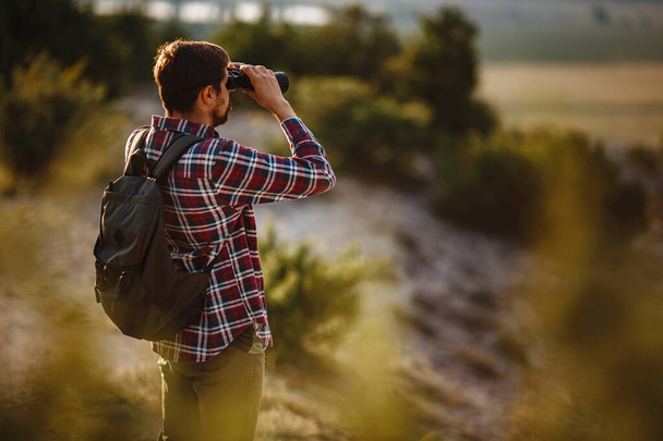 Kerl mit Ferngläsern in der Hand. Mann im T-Shirt mit Rucksack. Junger Kaukasier bei Wanderung in Tallandschaft - Foto, Bild