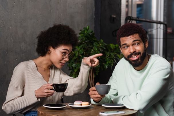 African american woman with coffee pointing with finger near smiling boyfriend in cafe  - 写真・画像