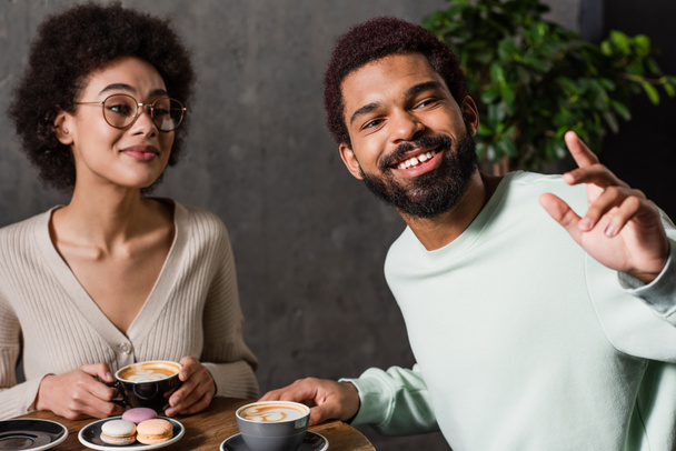 Smiling african american man looking away near girlfriend with coffee and macaroons in cafe  - Foto, Imagem