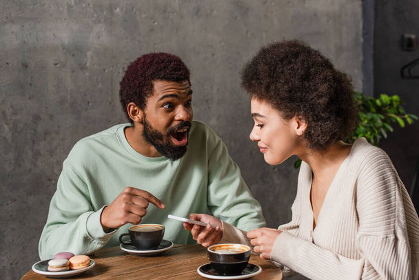 Excited african american man pointing with finger near girlfriend with smartphone and coffee in cafe  - Photo, Image