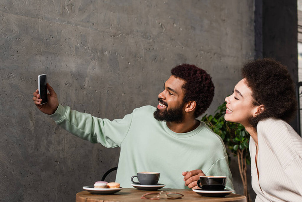 Vista laterale della coppia afro-americana sorridente che prende selfie vicino a caffè e macaron nel caffè  - Foto, immagini
