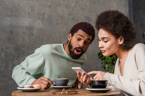 Homem americano africano incrível sentado perto da namorada com smartphone e café no café  - Foto, Imagem