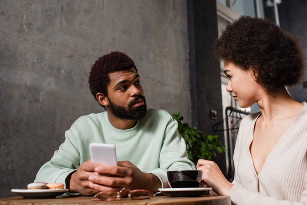 Afrykański Amerykanin ze smartfonem patrząc na dziewczynę przy kawie i rozmazane makaroniki w kawiarni  - Zdjęcie, obraz