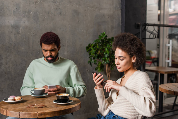 African american woman using smartphone near boyfriend and coffee in cafe - Фото, изображение