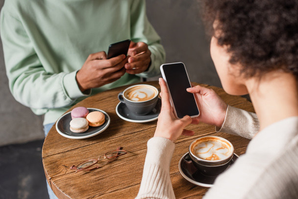 Smartphone in hands of african american woman near cappuccino and blurred boyfriend in cafe  - 写真・画像