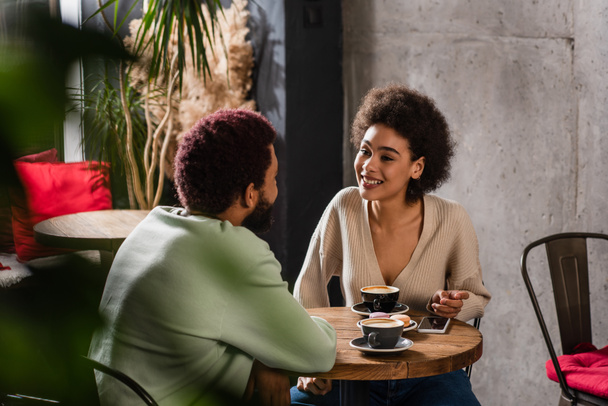 Smiling woman talking to african american boyfriend near coffee and smartphone  - Фото, зображення