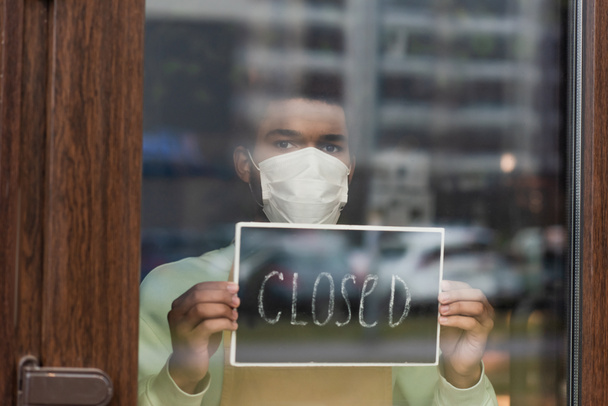 Afrykański amerykański barista w masce medycznej trzymając tablicę z zamkniętymi literami w kawiarni  - Zdjęcie, obraz