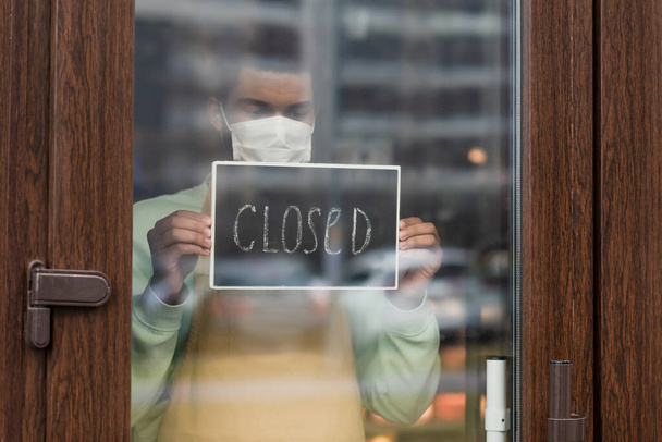 Barista afro-américaine en masque médical avec tableau noir avec lettrage fermé debout près de l'entrée dans le café  - Photo, image