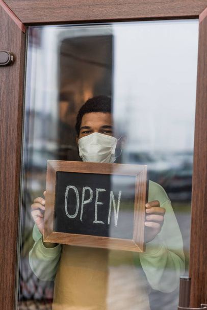 Tıbbi maskeli Afrikalı Amerikalı barista elinde kafenin kapısının yanında açık harflerle karatahta tutuyor.  - Fotoğraf, Görsel