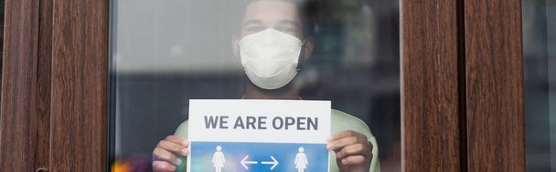 Afroamerikanischer Barista in medizinischer Maske hält Schild mit offenem Schriftzug in der Nähe der Cafétür, Banner  - Foto, Bild