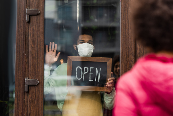 Tıbbi maskeli Afrikalı Amerikalı kahveci elinde açık harfli tabela ile bulanık müşterinin yanındaki kafede.  - Fotoğraf, Görsel