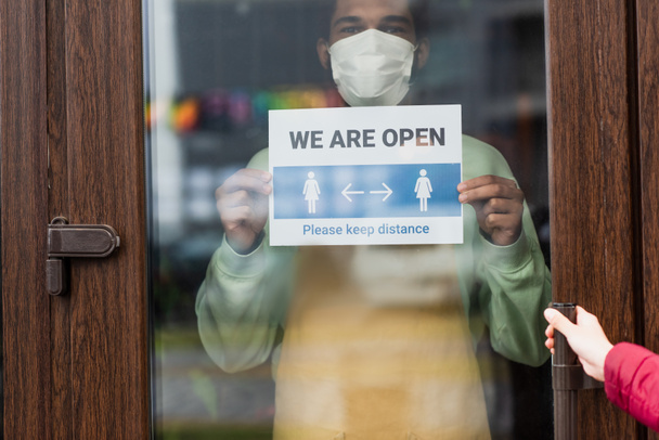 Αφρο-Αμερικανός barista σε ιατρική μάσκα κρατώντας πινακίδα με είμαστε ανοικτή επιστολόχαρτα κοντά στον πελάτη στην καφετέρια  - Φωτογραφία, εικόνα