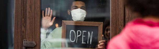 Barista afro-américaine dans un masque médical tenant un tableau noir avec lettrage ouvert et main agitant près du client flou dans un café, bannière  - Photo, image