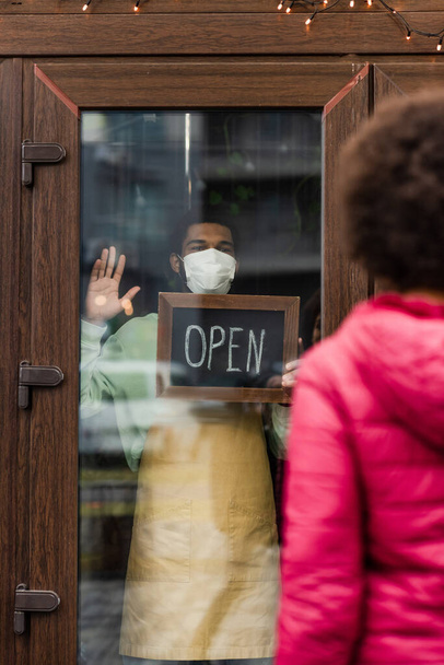 アフリカ系アメリカ人バリスタで医療用マスク手を振って、近くのカフェでオープンレタリングで黒板を保持  - 写真・画像