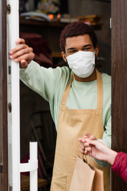 African american barista in medical mask giving paper bag to blurred client near door of cafe  - Фото, изображение