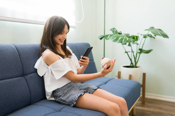 Chica joven y feliz tomando una foto con un teléfono inteligente a una hermosa magdalena deliciosa para publicar en sus redes sociales - Foto, imagen