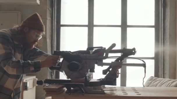 Mittlere Aufnahme eines kaukasischen Zimmermanns mit kariertem Hemd, Hut und Brille mit automatischer Kreissäge zum Schneiden von Brettern - Filmmaterial, Video