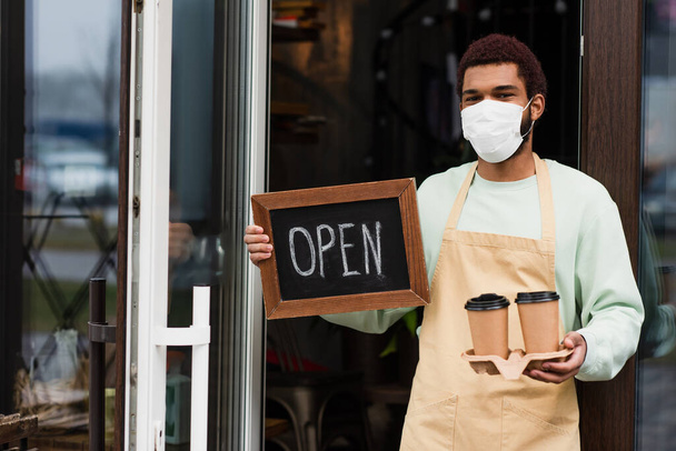 African american barista in medical mask holding coffee to go and chalkboard with open lettering near door of cafe  - Photo, Image