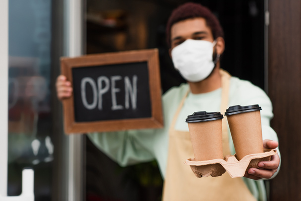 Coffee to go in hand of african american barista in medical mask holding chalkboard with open lettering on blurred background  - Photo, Image