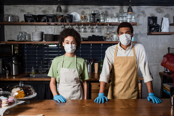 Afroamerikanische Baristas in Latexhandschuhen und medizinischen Masken stehen im Café  - Foto, Bild