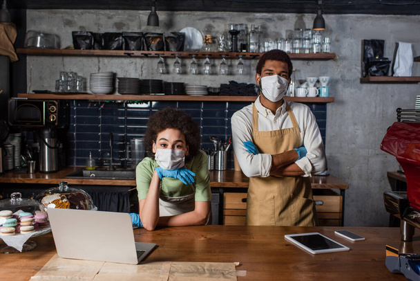 Afro-Amerikaanse barista in medisch masker en latex handschoenen staan in de buurt van apparaten en collega in cafe  - Foto, afbeelding