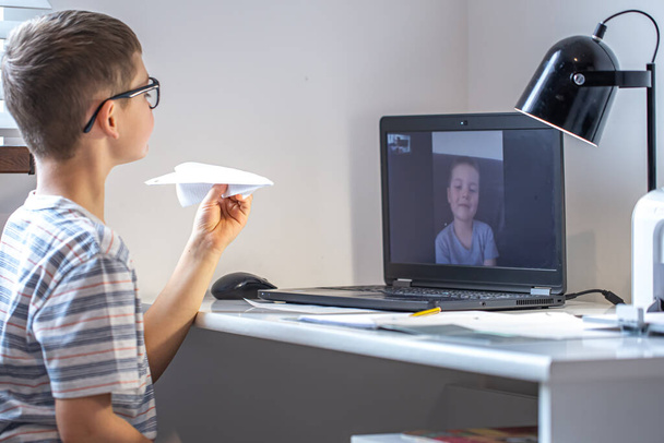 Ένας μαθητής δημοτικού σχολείου κάθεται σε ένα γραφείο μπροστά από ένα φορητό υπολογιστή και επικοινωνεί μέσω βίντεο σύνδεση στο διαδίκτυο στο σπίτι. - Φωτογραφία, εικόνα