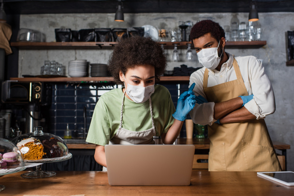African american barista in medical mask pointing at colleague near laptop in cafe  - Photo, Image