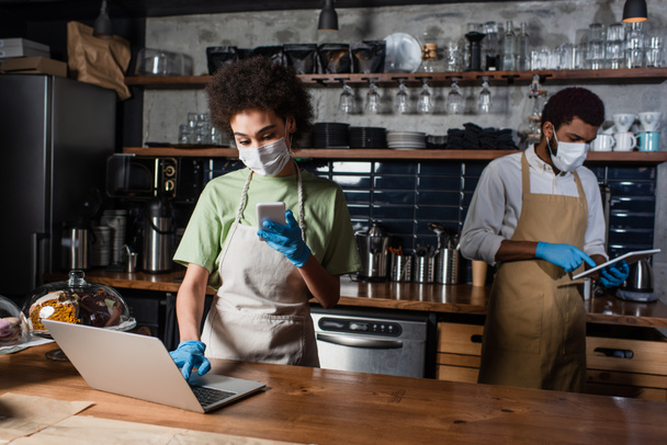 Africano americano barista em máscara médica usando smartphone e laptop perto colega no café - Foto, Imagem