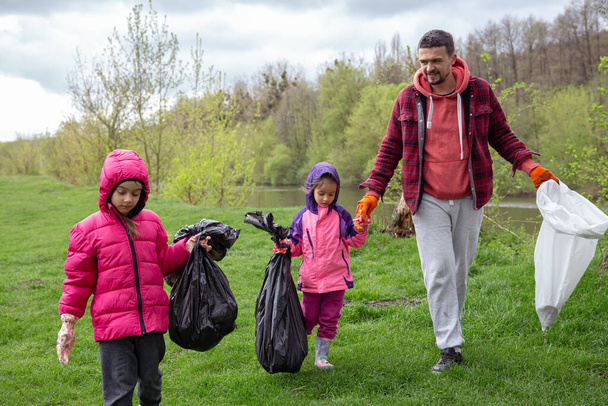 Zwei kleine Mädchen mit ihrem Vater, die mit Müllsäcken in der Natur unterwegs sind, säubern die Umwelt. - Foto, Bild