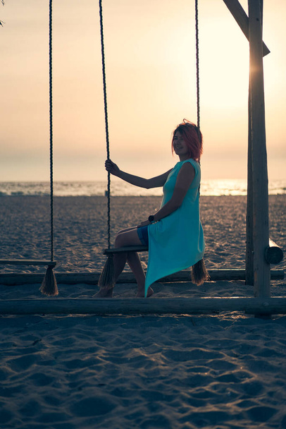 Młoda atrakcyjna kobieta z różowymi włosami spoczywa na huśtawce na plaży o zachodzie słońca. - Zdjęcie, obraz