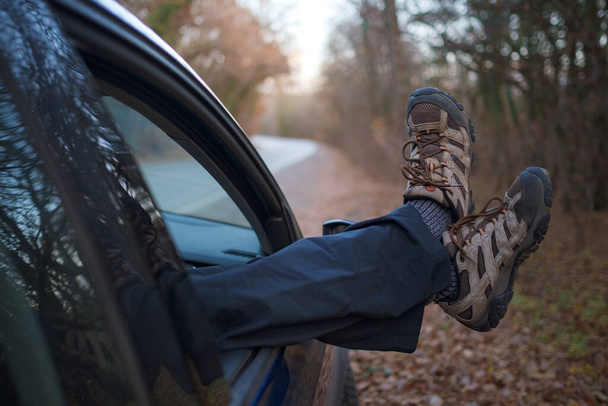 pies masculinos en botas de trekking que sobresalen por la ventana del coche. Viaje en coche de otoño al atardecer en el bosque. El concepto de libertad de movimiento. Conducir a través de un fin de semana - Foto, imagen