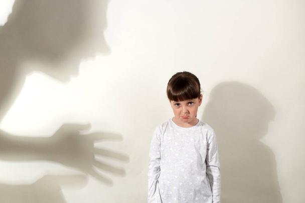 Shadow of aggressive human hits little girl, kid suffering from domestic violence, shadow silhouette on the wall to represent abuse, fear, or bully, offended child looks at camera with pout lips. - Zdjęcie, obraz